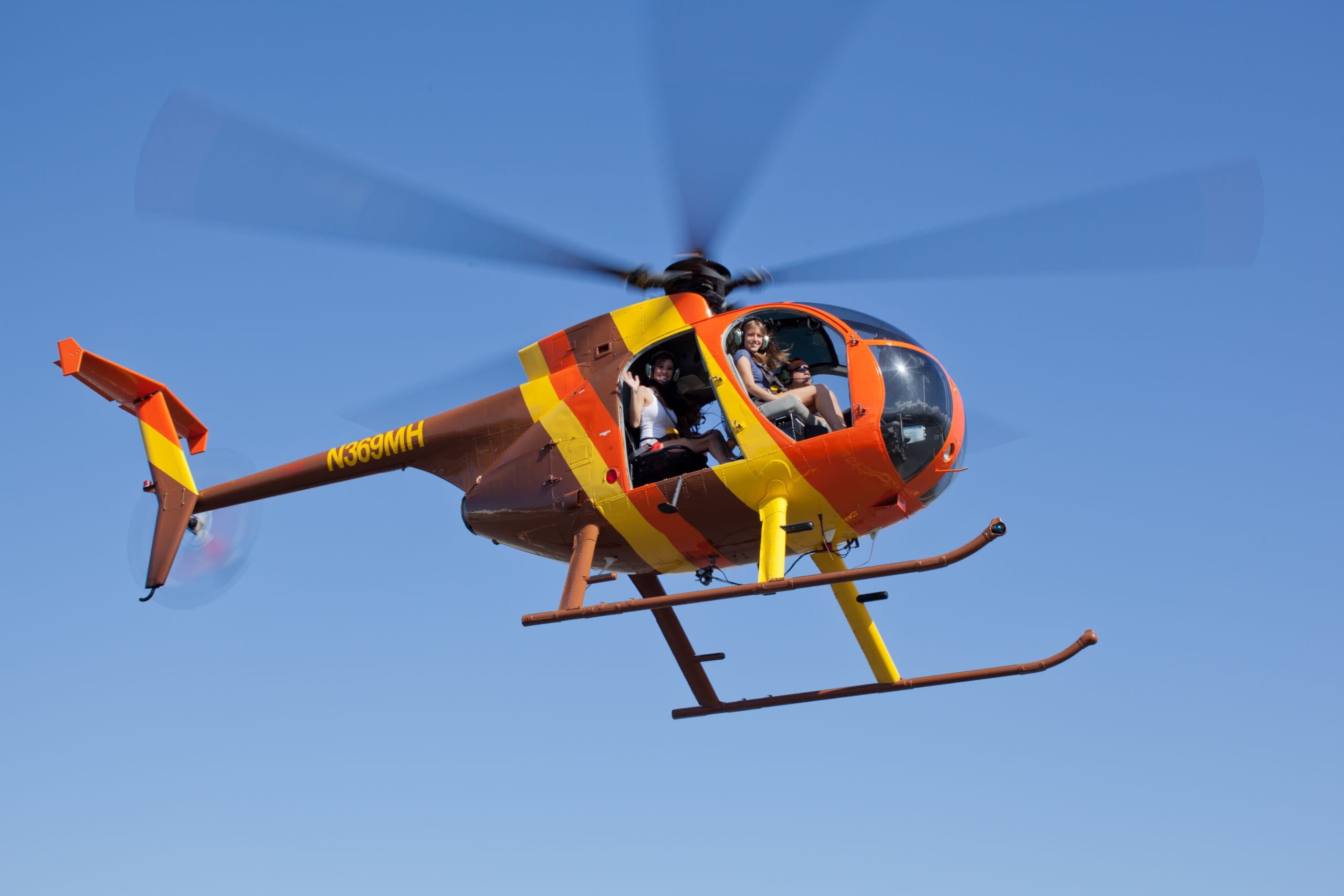 Doors-off Hughes 500D helicopter in flight against a blue sky with passengers aboard